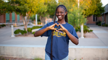 Fall 2024 graduate Mimi Owolabi wears a Phoenix College t-shirt and holds her hands in a heart shape while standing on campus