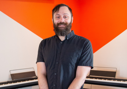 Phoenix College alumnus Jason Mohlenbrock stands in front of two pianos against an orange wall in his youth piano academy, named Blue in Green after his favorite Miles Davis tune. 